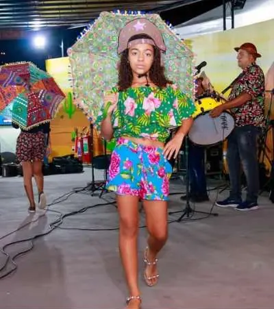 Jovem desfila com roupa colorida e chapéu de flores diante de uma banda