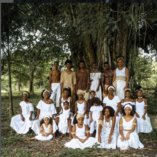 Grupo de crianças, jovens e adultos vestidos de branco embaixo de uma grande árvore