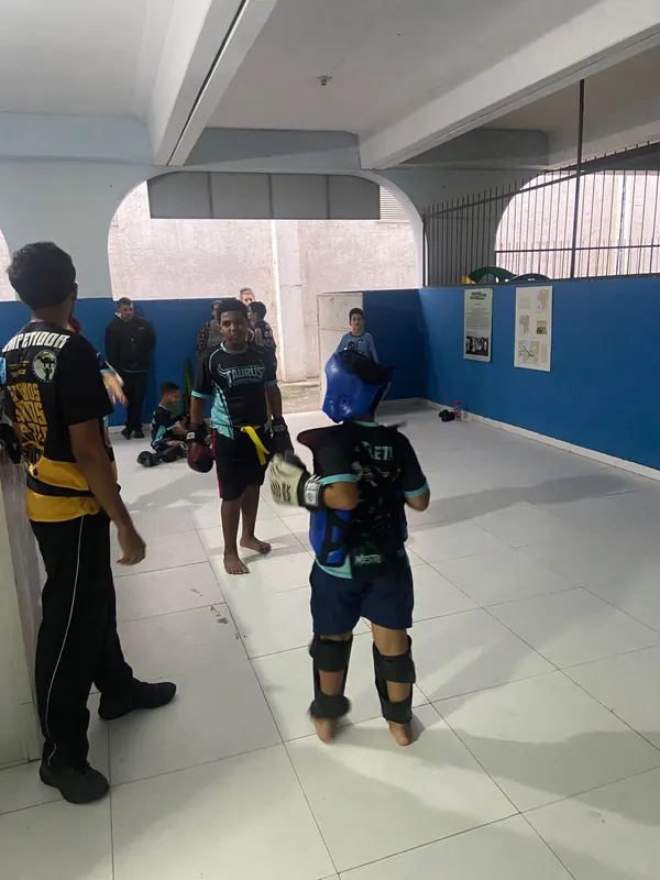 Criança com capacete e protetores azuis treina com luvas no salão do espaço, observada pelo instrutor