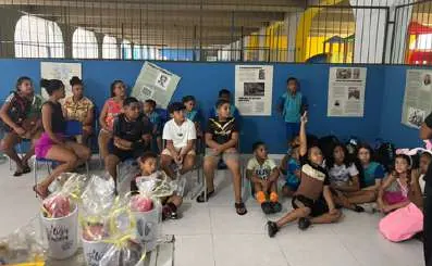 Crianças sentadas no salão de paredes azuis, com kits de presente embalados à frente