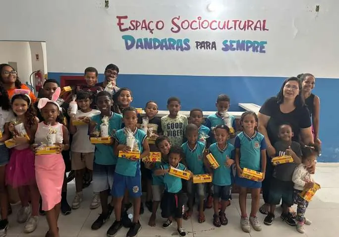 Crianças, algumas com orelhas de coelho, seguram caixas de bombom em frente ao letreiro do espaço